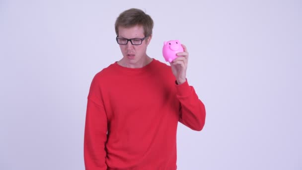 Stressed young man holding piggy bank and giving thumbs down - Footage, Video