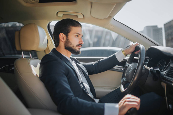Handsome and confident young man sits in car and looks straight forward. He drives car. It looks luxury inside. He holds one hand on steering wheel and another one on handbrake. - Foto, imagen
