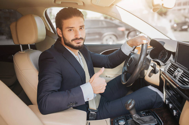 Nice picture of young businessman sitting in luxury car and holding big thumb up. He hold one hand on steering wheel. Young man recommends this car. - Photo, Image