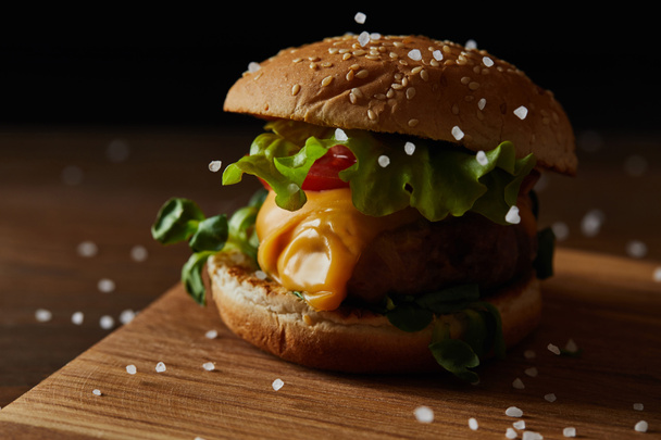 close up of salt falling on tasty meat burger with cheese and greenery on wooden chopping board - Foto, imagen