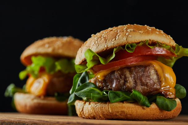 selective focus of delicious meat burgers with cheese and vegetables on wooden chopping board isolated on black - Foto, Imagem