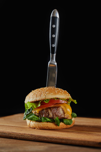 knife in delicious meat burger with cheese, greenery and tomatoes and sesame isolated on black - Fotografie, Obrázek