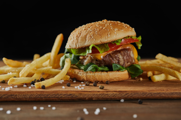 golden french fries with tasty meat burger with scattered salt and black pepper on wooden chopping board isolated on black - Photo, Image
