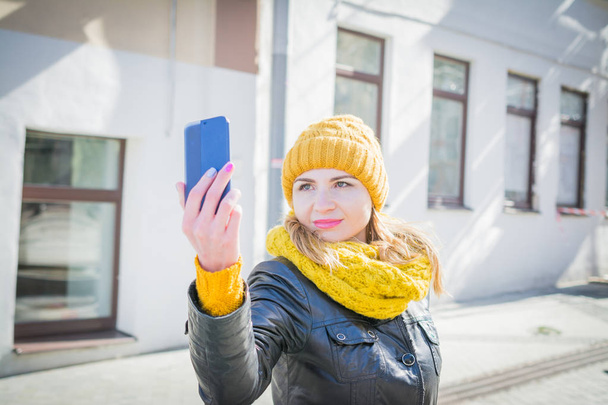 Beautiful woman in a yellow knitted hat takes pictures of herself on her mobile phone - Φωτογραφία, εικόνα