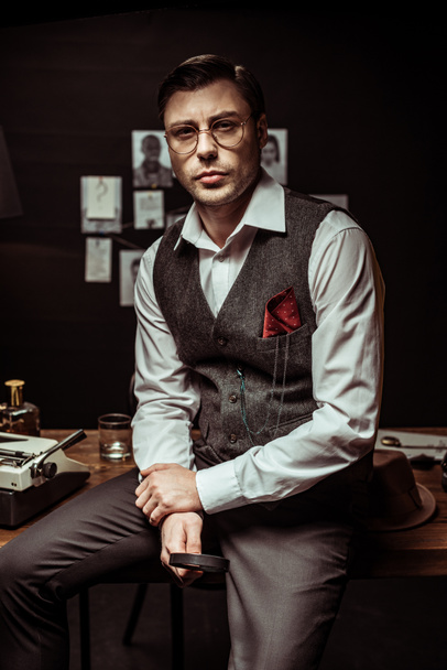 Detetive em óculos sentado na mesa e segurando lupa no escritório escuro
 - Foto, Imagem