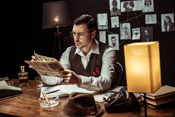 Concentrated detective in glasses reading newspaper in dark office - Zdjęcie, obraz