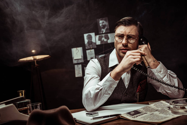 Pensive detective in glasses talking on telephone in dark office - 写真・画像