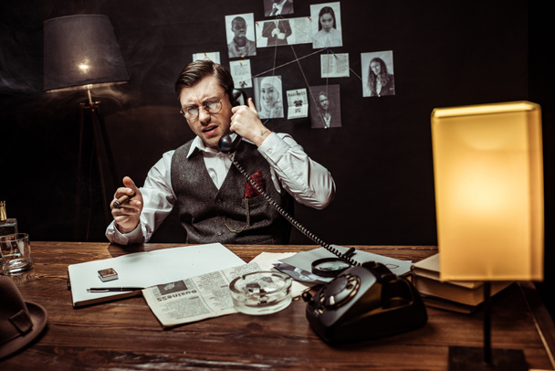Роздратований детектив в окулярах тримає сигару і розмовляє по телефону в темному офісі
 - Фото, зображення