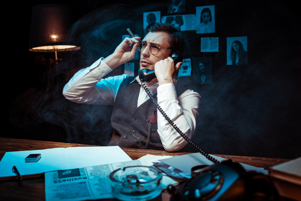 Detektiv mit Brille hält Zigarre in der Hand und telefoniert im dunklen Büro - Foto, Bild