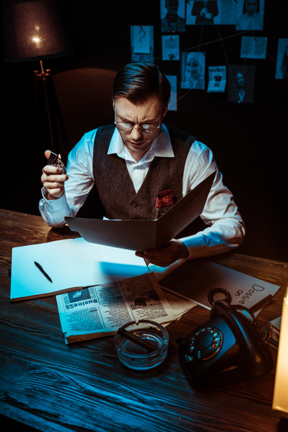 Vista de ángulo alto del detective concentrado en gafas que lee el expediente en la oficina oscura
 - Foto, Imagen