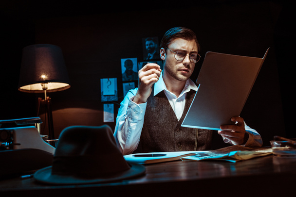 Концентрований детектив в окулярах читає досьє в темному офісі
 - Фото, зображення