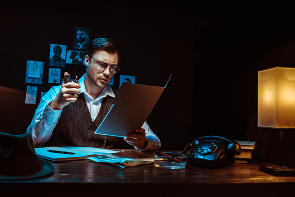Συγκεντρωμένη ντετέκτιβ σε γυαλιά διαβάζοντας φάκελο σε σκοτεινό γραφείο - Φωτογραφία, εικόνα