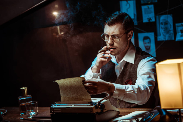 Detective in glasses smoking cigar while using typewriter in dark office - Foto, imagen