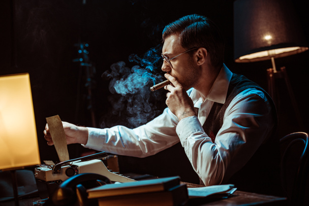 Detetive em óculos fumar charuto ao usar máquina de escrever no escritório escuro
 - Foto, Imagem