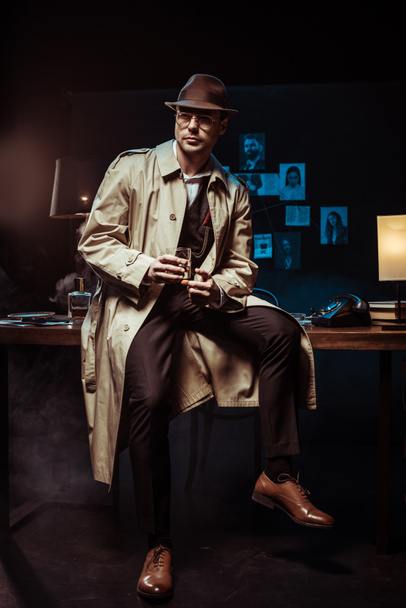 Detective in hat and trench coat sitting on table with glass of cognac - Foto, imagen