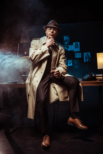 Detective in trench coat and hat sitting on table and holding glass of cognac - Photo, image