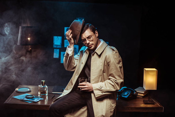 Pensive detective putting on hat while holding glass of cognac - 写真・画像