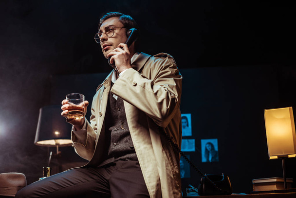 Low angle view of detective in glasses talking on telephone and holding glass of cognac - Foto, Imagem