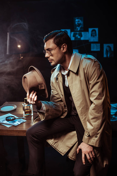 Pensive detective in glasses and trench coat sitting on table and holding hat - Фото, зображення