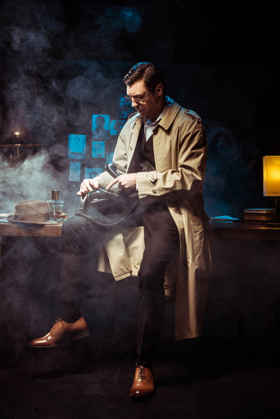 Detective in trench coat sitting on table and using telephone in dark office - Zdjęcie, obraz
