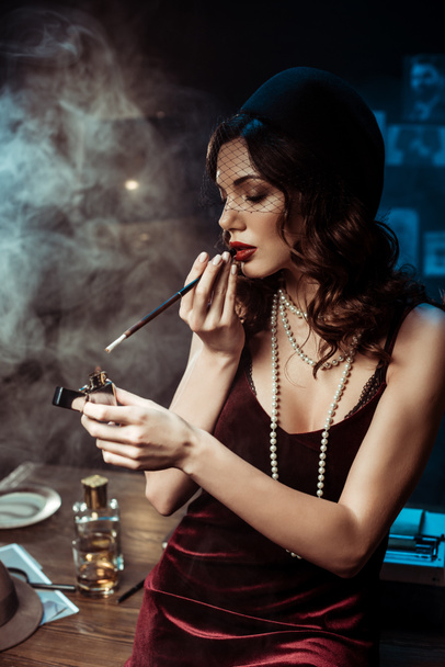 Beautiful woman with mouthpiece lighting cigarette with lighter in dark office - Photo, image