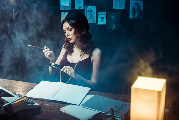 attraktive Frau mit Fascinator-Hut mit Mundstück und Lupe im dunklen Büro - Foto, Bild