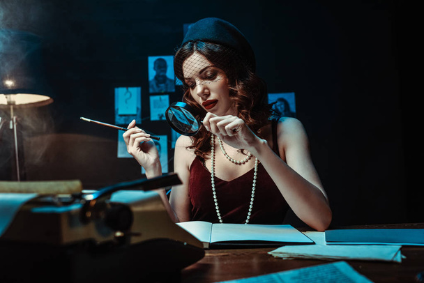 Attractive woman in fascinator hat holding mouthpiece and magnifier in dark office - Fotó, kép