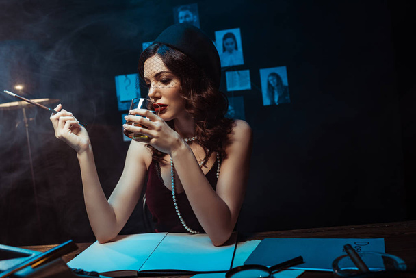 attraktive Frau mit Fascinator-Hut mit Mundstück und Glas Cognac im dunklen Büro - Foto, Bild