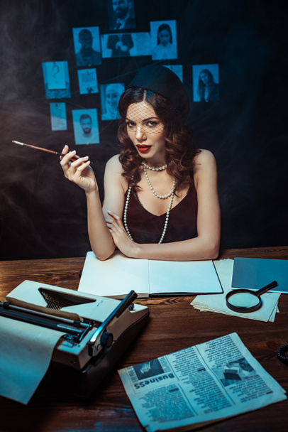 Mujer bonita en vestido con boquilla sentada en la mesa con máquina de escribir en la oficina oscura
 - Foto, Imagen