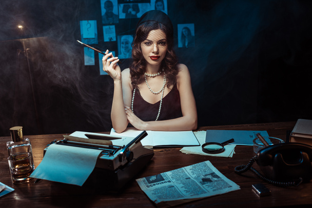 Mujer bonita en vestido con boquilla sentada en la mesa con máquina de escribir en la oficina oscura
 - Foto, Imagen