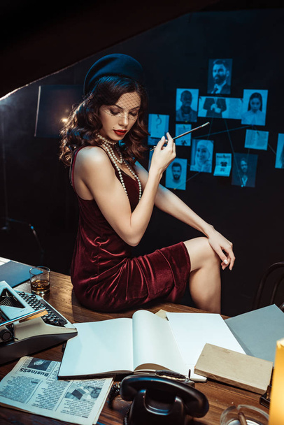 Mujer pensativa en vestido y sombrero fascinador sosteniendo boquilla y mirando documentos en la oficina oscura
 - Foto, Imagen