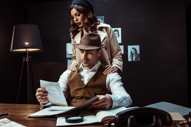 Detective and beautiful woman in trench coat looking at photo in dark office - Foto, immagini