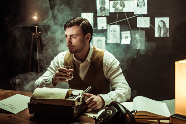 Detective sosteniendo un vaso de coñac mientras usa la máquina de escribir en la oficina oscura
 - Foto, Imagen