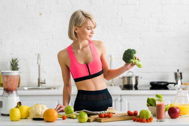 attractive blonde girl in sportswear holding green broccoli in kitchen  - Фото, зображення