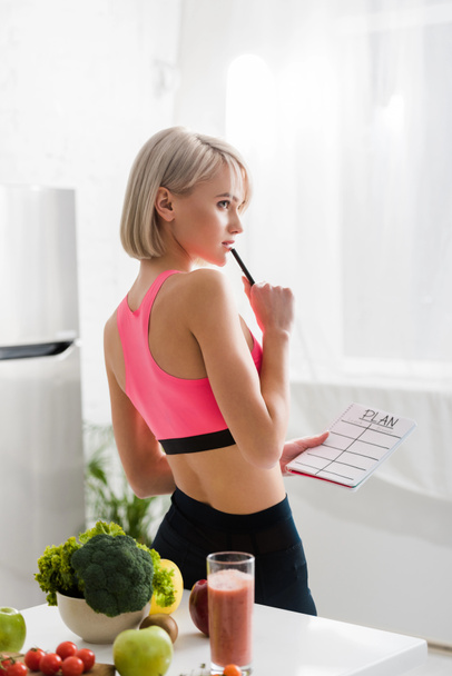 pensive blonde woman in sportswear holding notebook with plan lettering in kitchen  - Photo, Image