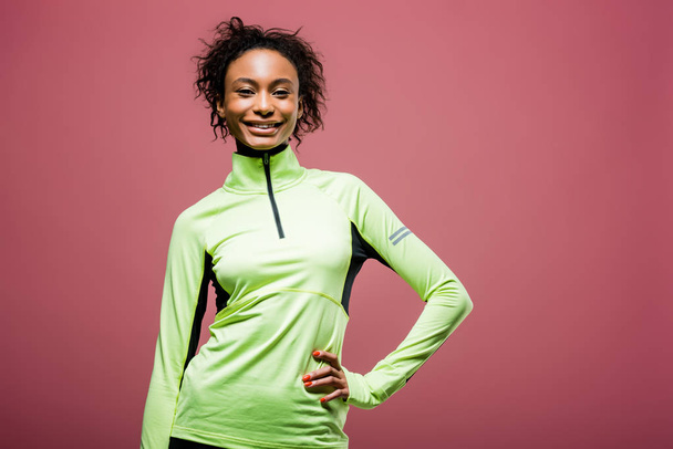 bela esportista afro-americana em jaqueta de pista olhando para a câmera e sorrindo isolado em rosa
 - Foto, Imagem