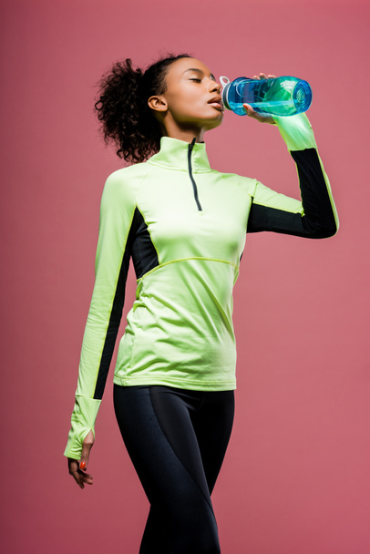 beautiful african american sportswoman drinking water from sport bottle isolated on brown - Photo, Image