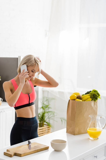 blonde girl in sportswear talking on smartphone near paper bag with groceries - Photo, Image