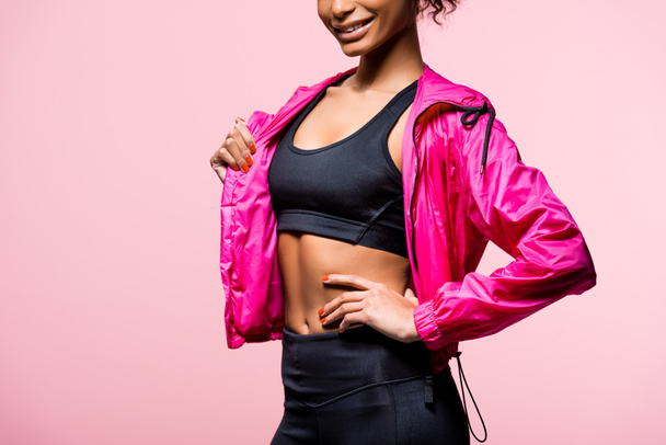 обрезанный вид африканской улыбающейся американской спортсменки в ветровке позирующей изолированной на розовом
 - Фото, изображение