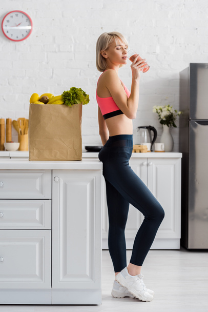 attractive blonde woman in sportswear drinking smoothie near paper bag with groceries - Фото, изображение