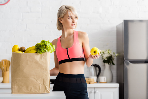 attractive blonde woman in sportswear holding apple near paper bag with groceries - Photo, Image