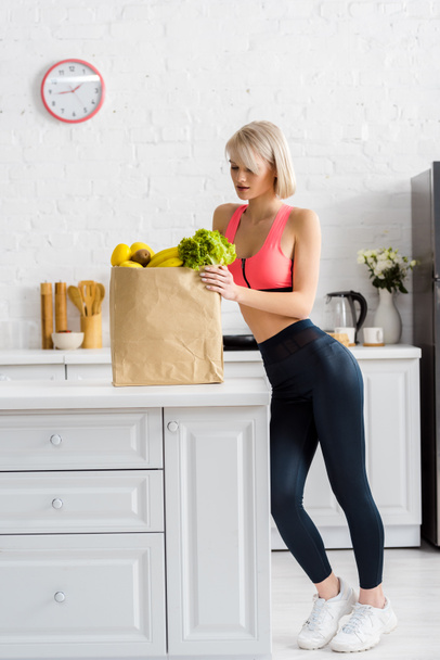 attractive blonde woman in sportswear looking at paper bag with groceries - Fotó, kép