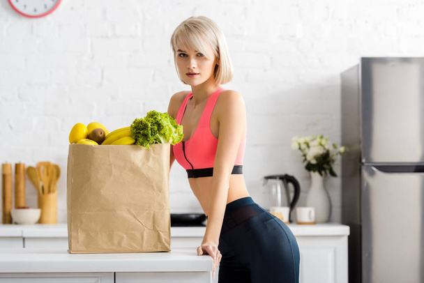 blonde woman in sportswear looking at camera near paper bag with groceries - Zdjęcie, obraz