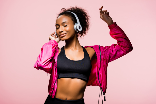 音楽を聴くと、ピンクに分離されたジェスチャーのヘッドフォンで美しいアフリカ系アメリカ人のスポーツウーマン - 写真・画像