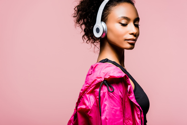 beautiful african american sportswoman in windbreaker and headphones isolated on pink with copy space - Photo, Image