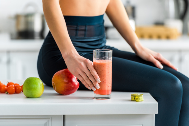 cropped view of woman holding glass of smoothie while sitting in kitchen near apples - Foto, Imagen