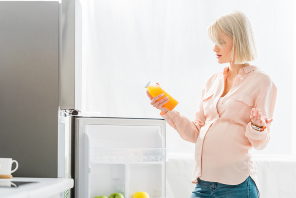 mujer embarazada rubia sosteniendo botella con jugo de naranja cerca de la nevera en la cocina
  - Foto, Imagen