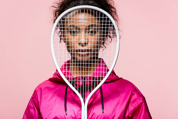 bela esportista afro-americana olhando para a câmera enquanto cobre o rosto com raquete de tênis isolado em rosa
 - Foto, Imagem