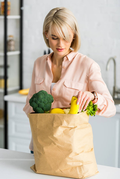 atractiva mujer rubia y embarazada mirando bolsa de papel con comestibles
  - Foto, imagen