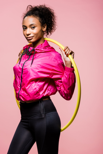 beautiful smiling african american sportswoman posing with hula hoop isolated on pink - Фото, зображення
